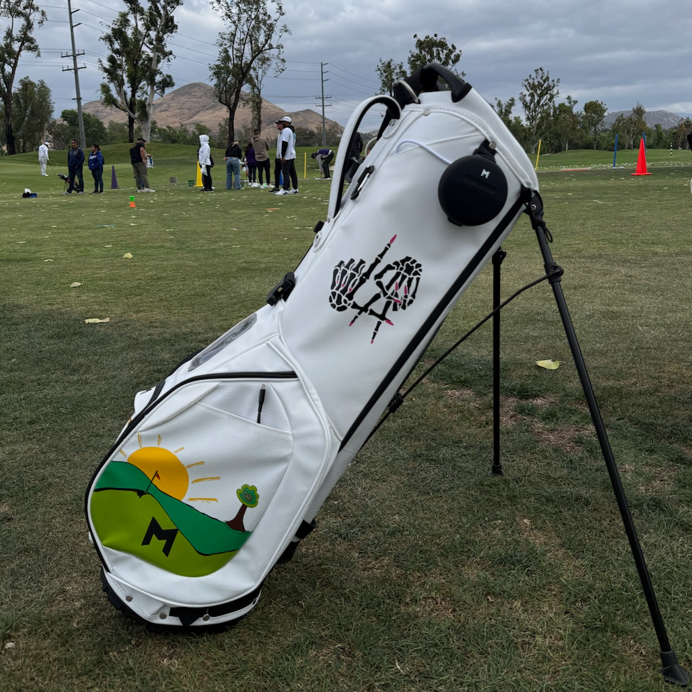 White golf bag with a colorful logo on a grassy field with people and trees in the background