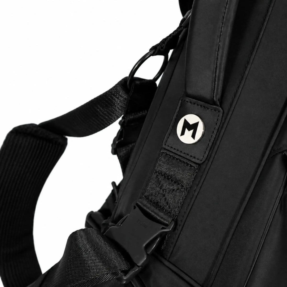 Close-up of a black backpack strap with a silver circular metal ball marker featuring a stylized letter 'M' on a white background.