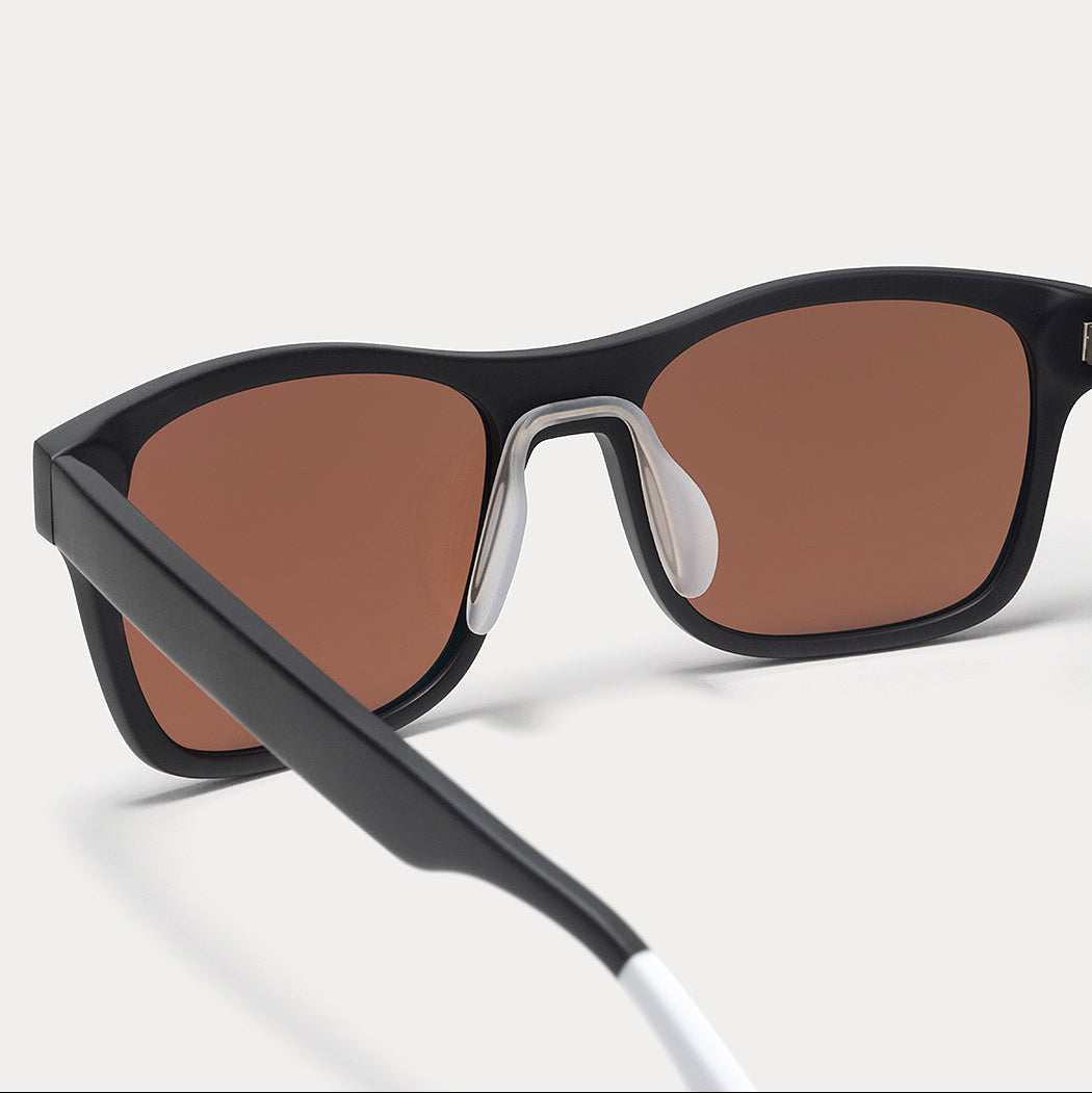 Close-up of a pair of sunglasses with a matte black frame, brown tinted lenses, and a white temple tip, shown on a light gray background.