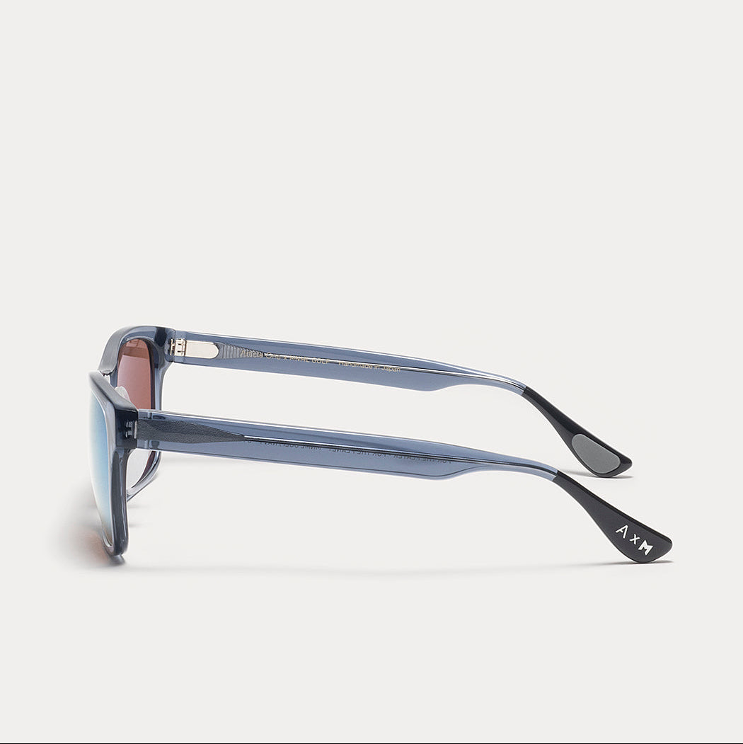Side profile of square-frame sunglasses with a translucent slate blue acetate frame, dark lenses, and a small white AXN logo on the temple tip, set against a light gray background.