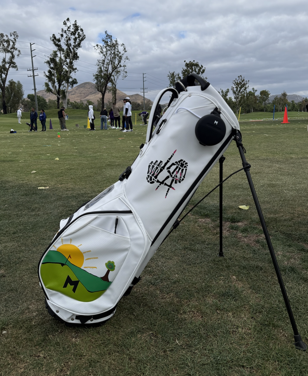 White golf bag with a colorful logo on a grassy field with people and trees in the background