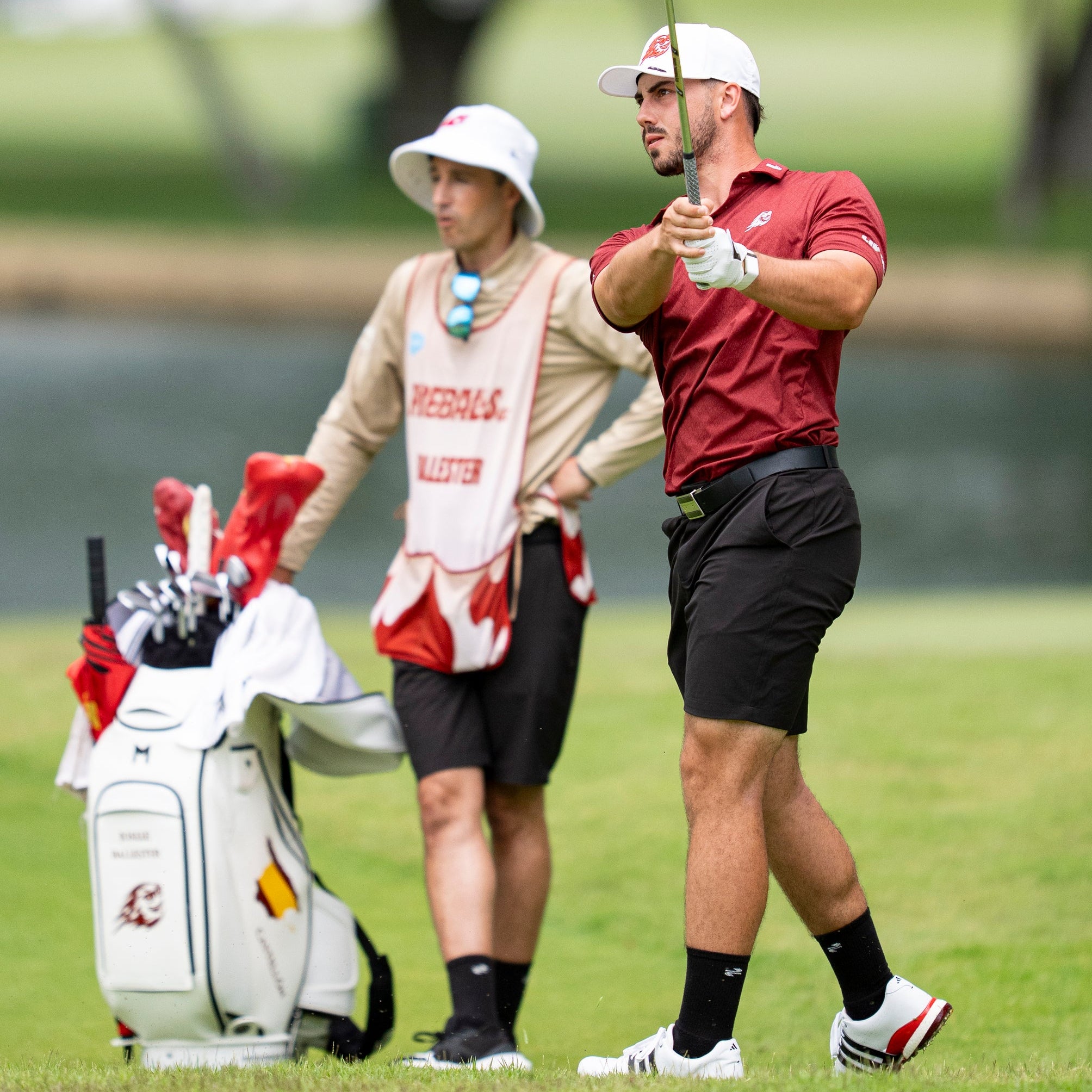 Golfer in red shirt and black shorts preparing to hit a golf ball with a caddie in the background.