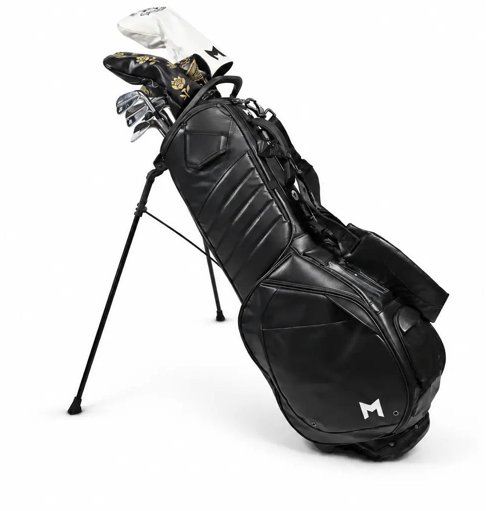A black stand golf bag with 14 golf clubs inserted, photographed on a clean white studio background. A small white stylized 'M' logo is visible near the lower front of the bag.
