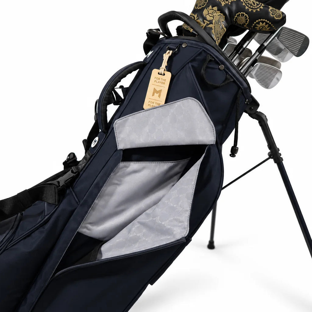 A navy blue stand golf bag with a light gray interior lining, shown with golf clubs inserted and a hanging wooden tag on a white background.