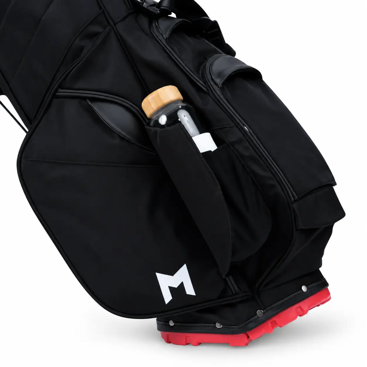 A close-up of a black golf bag with a white stylized 'M' logo near the bottom front pocket and a bright red outsole. A clear water bottle with a light wood cap is tucked into a side pocket.