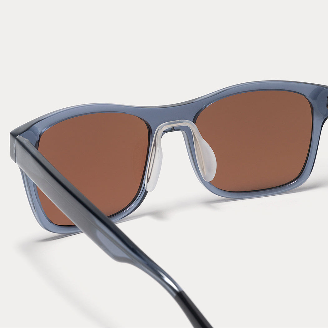 Close-up of a pair of sunglasses with a translucent slate blue frame and warm brown lenses, shown on a light gray background.