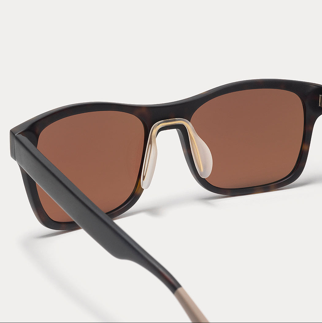 Close-up of a pair of sunglasses with a dark tortoiseshell frame, brown lenses, and a gold metal bridge, shown on a light gray background.