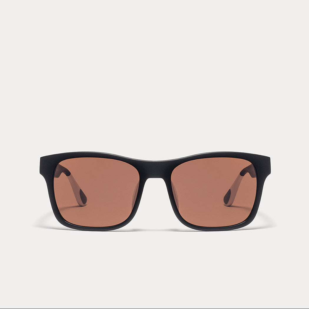 A pair of square sunglasses with a thick black frame and warm brown-tinted lenses, shown against a light gray background.