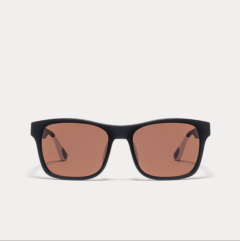 A pair of square sunglasses with a thick black frame and warm brown-tinted lenses, shown against a light gray background.