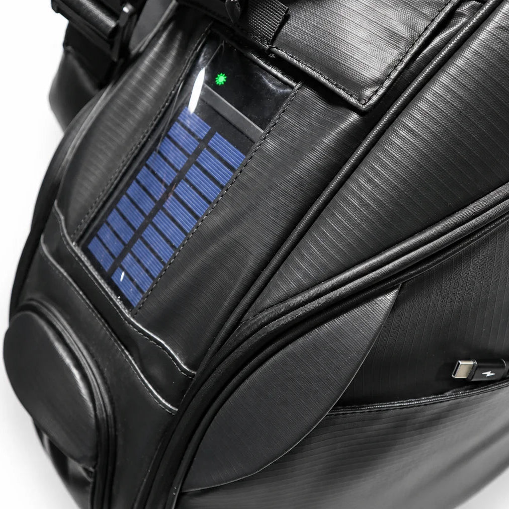 Close-up of a black textured golf bag featuring a built-in solar panel on the front pocket, with a small green LED light illuminated, set against a plain white background.