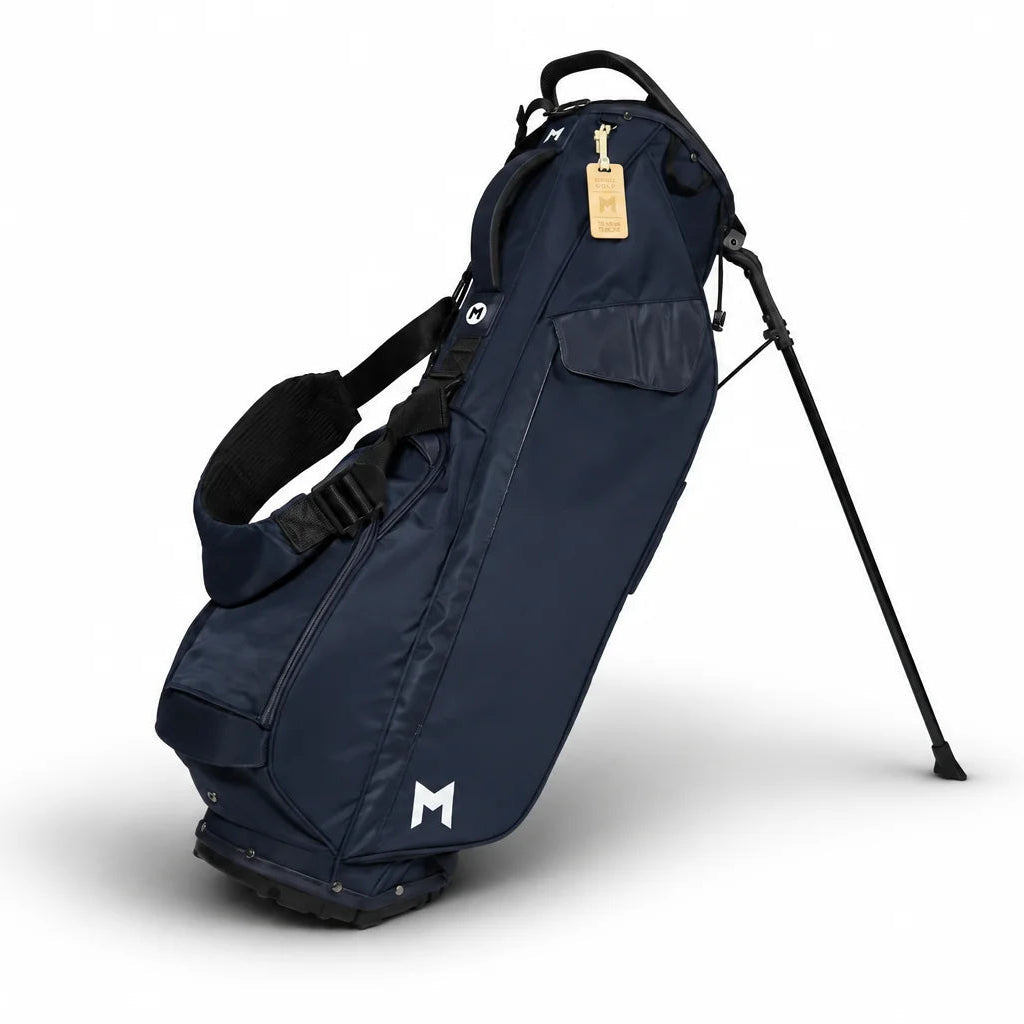 A navy blue stand golf bag with black legs and straps, featuring a white stylized 'M' logo near the bottom and a small gold hangtag, shown on a white background.