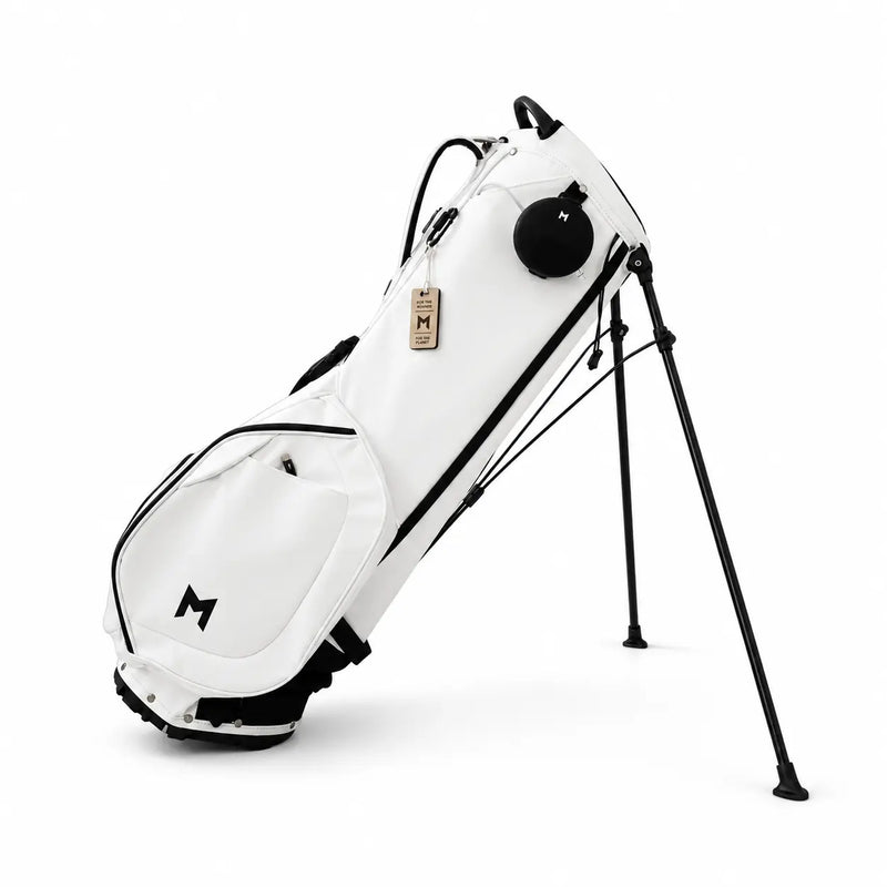 A white stand golf bag with black accents, including a black tripod-style leg system, black trim, and a black logo on the lower front pocket. It features a small tan leather hangtag with an 'M' logo.