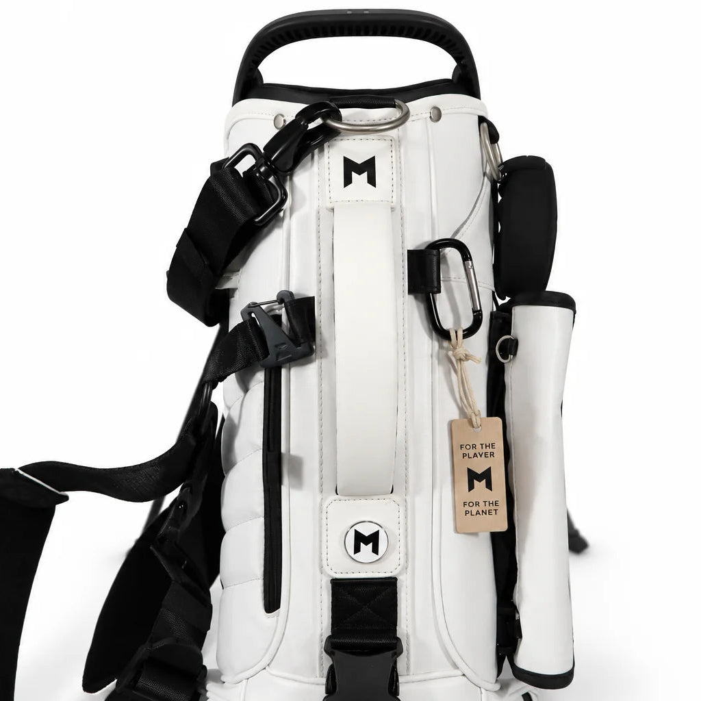A white and black golf bag shown from the back, featuring multiple magnetic pockets, black straps, a top carry handle, and a hanging tag with the M-Head logo.